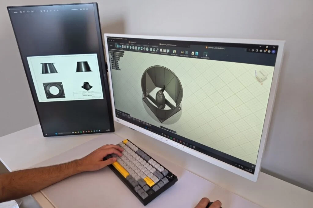 Dos monitores del taller con el modelo 3D de una pieza abierto en el programa de diseno, y las manos sobre el teclado.