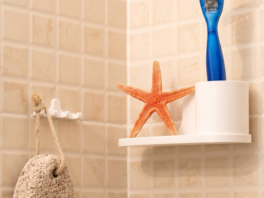 Estante blanco de esquina con una maquinilla y una estrella de mar, y un colgador con una piedra pómez, sobre azulejo beige.