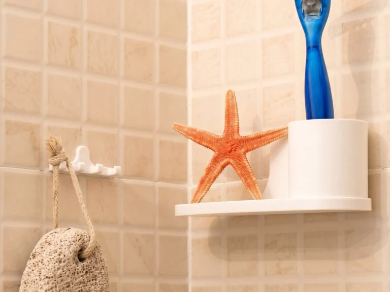 Estante blanco de esquina con una maquinilla y una estrella de mar, y un colgador con una piedra pómez, sobre azulejo beige.