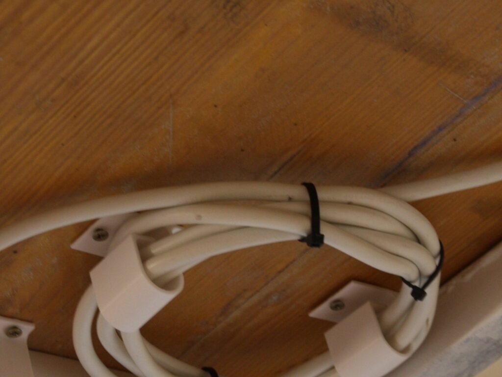 Pinzas blancas impresas en 3D atornilladas bajo un tablero de madera, sujetando un cable enrollado y una regleta.