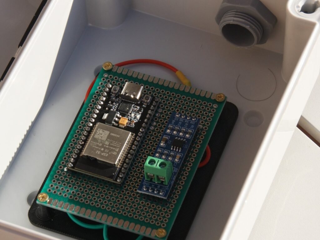 Placa electrónica verde montada sobre una bandeja negra impresa en 3D, dentro de una caja estanca blanca.