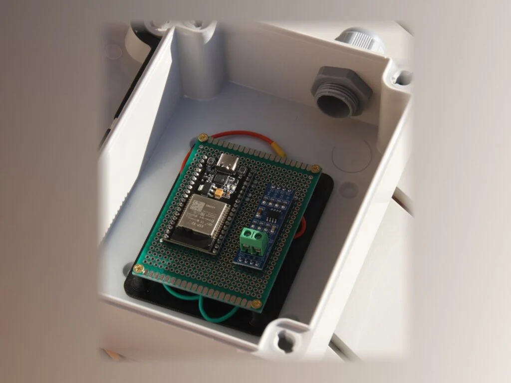 Placa electrónica verde montada sobre una bandeja negra impresa en 3D, dentro de una caja estanca blanca.