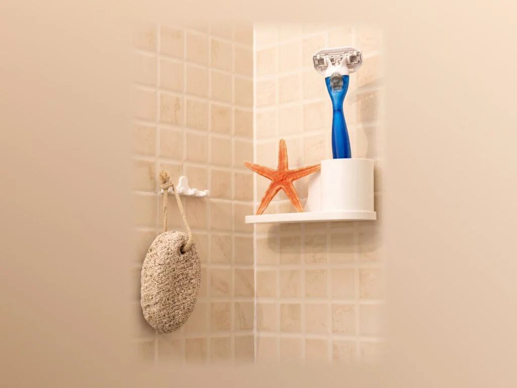 Estante blanco de esquina con una maquinilla y una estrella de mar, y un colgador con una piedra pómez, sobre azulejo beige.