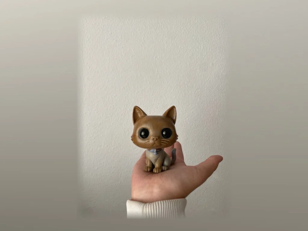 Figura de un gato marrón con los ojos grandes, sostenida en la palma de una mano.